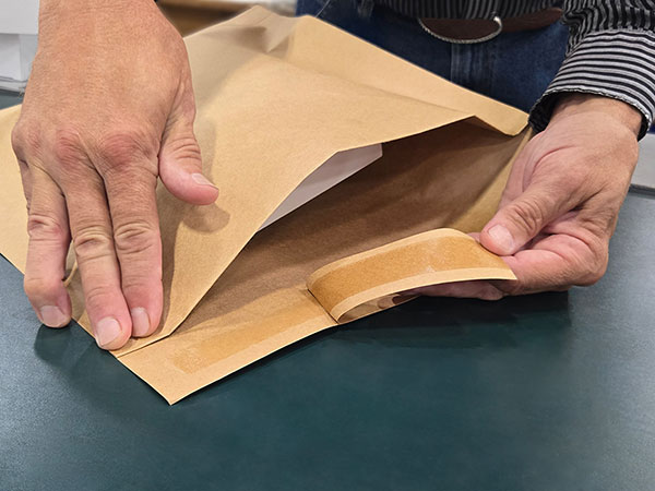 Hands peeling a adhesive strip to seal a brown paper mailer envelope.