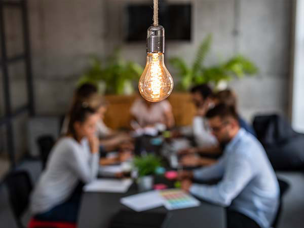 Light bulb brightly lit in the foreground, with a team of workers in the background meeting at a conference table
