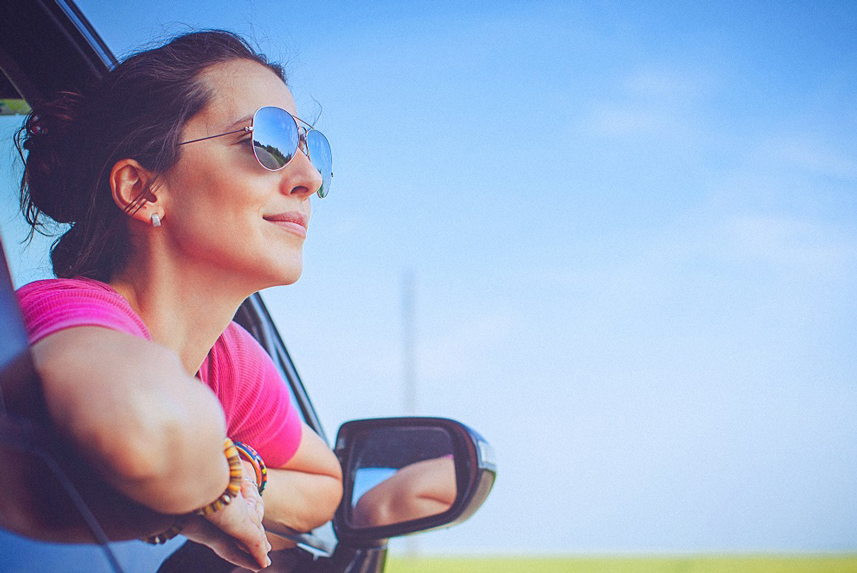 Woman in pink shirt and sunglasses, leaning out car window and smiling with blue sky in the background