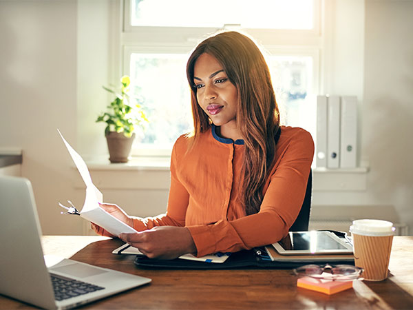 Woman at desk with laptop open and reviewing paper documents