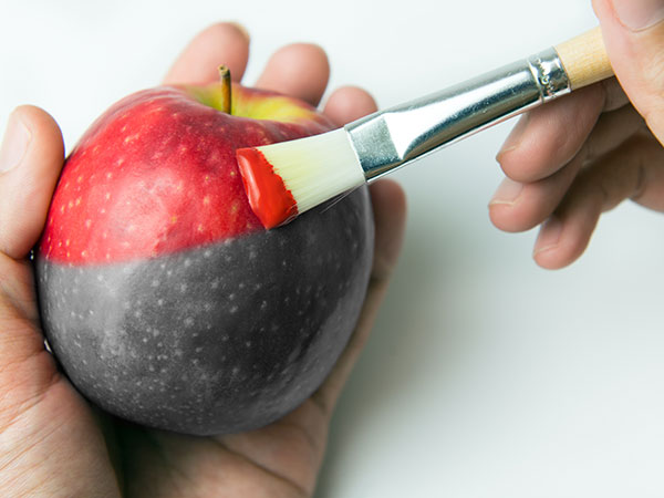Hand holding a grayscale apple while another is holding a paintbrush and painting the apple red