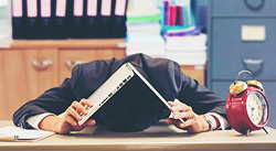 Photo of a man slumped on a desk with an open laptop covering his head
