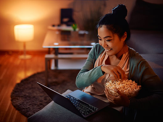 Smiling Asian woman eats popcorn while watching something on her laptop