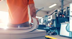 Man in orange polo shirt and blue jeans picking up a stack of paper, large commercial printing is in the background