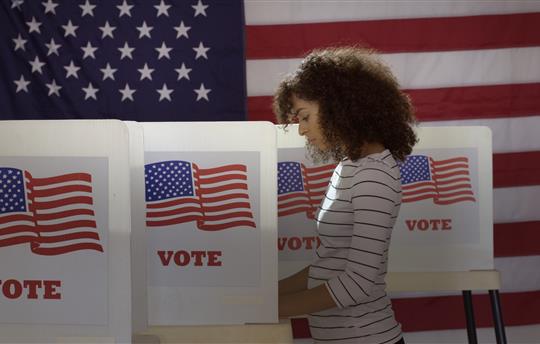 Profile, medium shot, young woman in polling station, voting in a booth with US flag in back