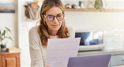 woman wearing glasses and looking at a printed document while sitting near her laptop