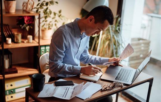 man in blue button down shirt sitting in home office paying bills on laptop