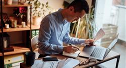 man in blue button down shirt sitting in home office paying bills on laptop