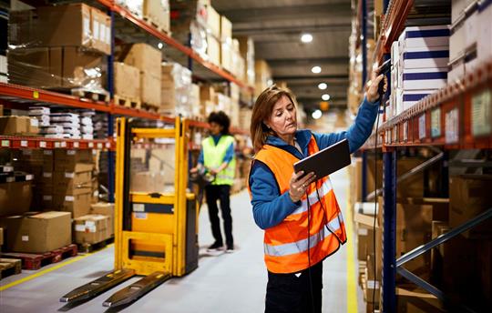 one female warehouse worker in bright yellow vest operating forklift as another female warehouse worker in a bright orange vest scans a label in a box