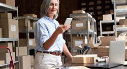 Female small business owner using mobile app checking parcel box and postal shipping order in warehouse
