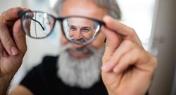 Older man with gray beard holding his eyeglasses out away from his face