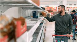 man shopping in large store, pushing a cart and looking at shelves with very little stock