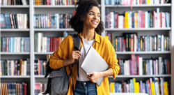 Portrait of a female student standing against background of bookshelves in university library holding laptop and backpack looking to the side
