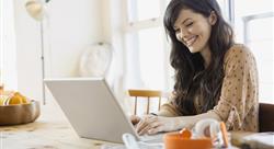 woman sitting at her dining room table, smiling while she views something on her laptop