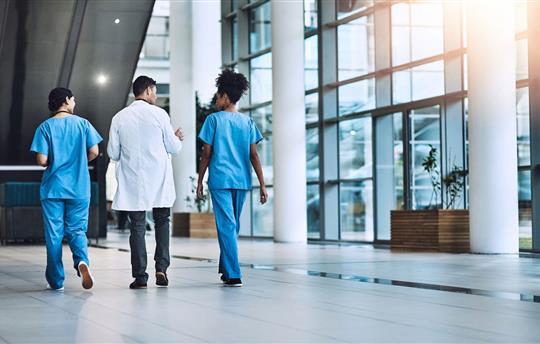 two healthcare workers in blue scrubs and one in a white coat walking and talking together through hospital lobby