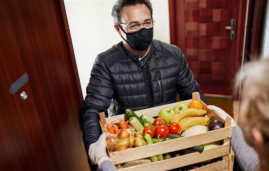 Man in black reusable face mask delivering crate of vegetables to elderly woman