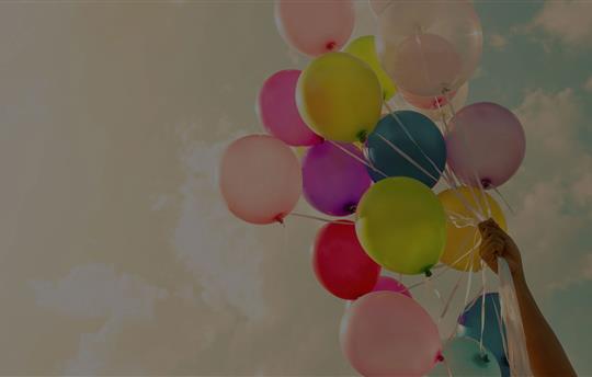 arm reaching up holding a bunch of multi-color balloons with the sky in the background